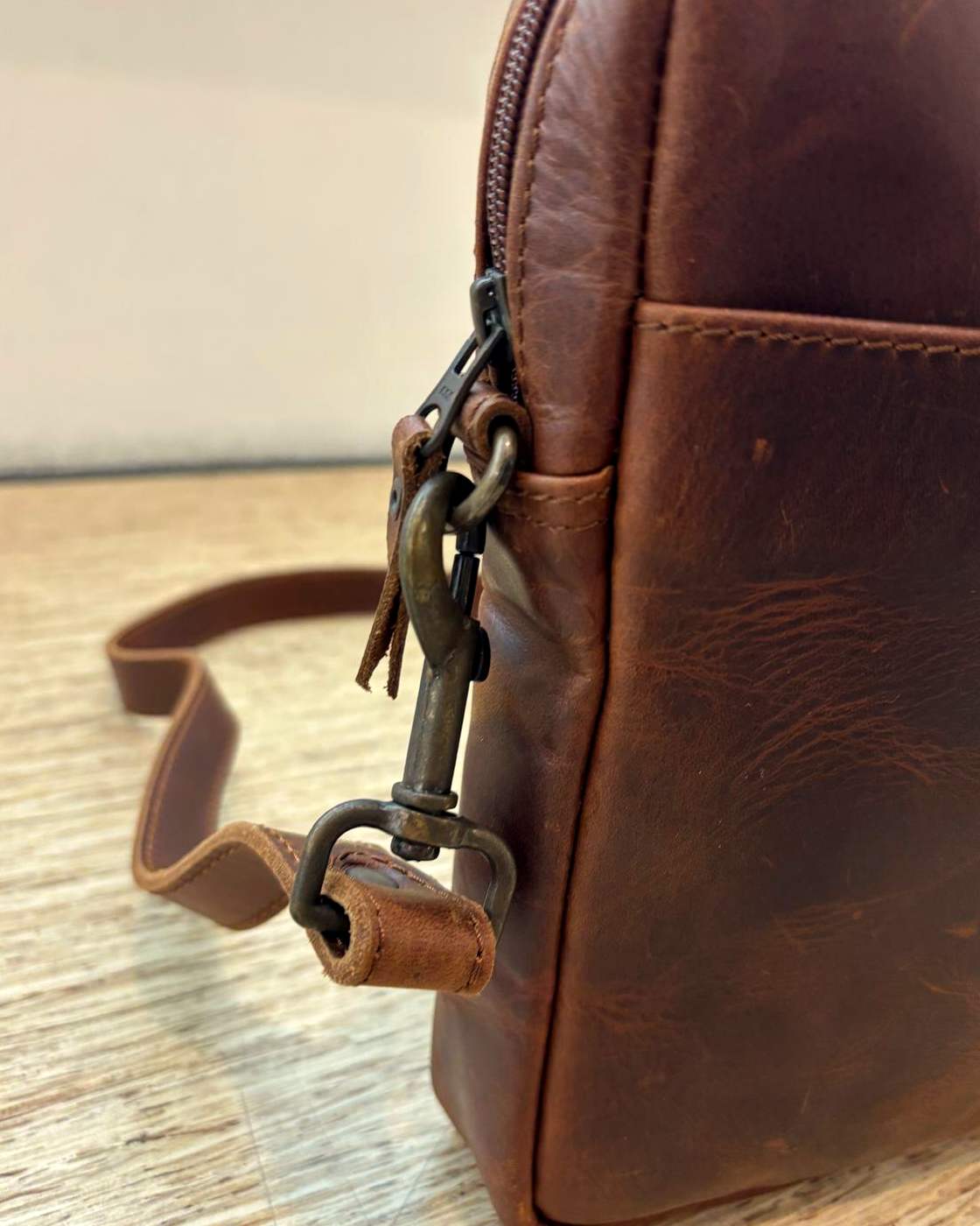 Close-up real photo of the YYZ Satchel strap hardware: a swivel clip hooked through a D-ring at the upper side seam of the bag, beside the main zipper end and leather pull tab.