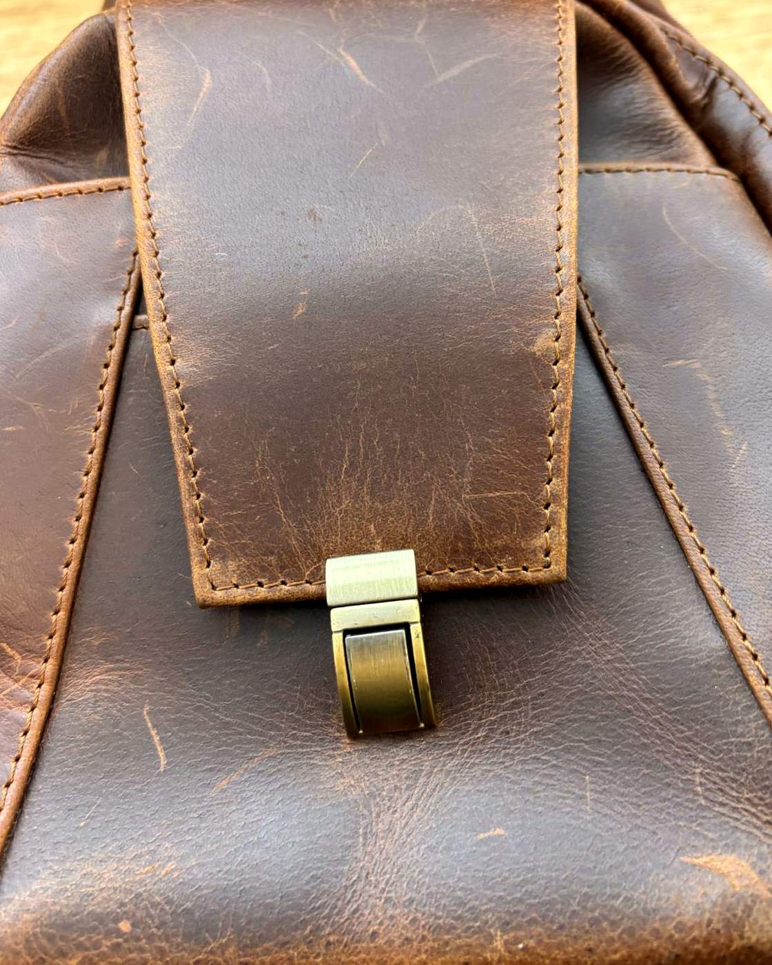 Close-up real photo of the Courier Sling front flap and its metal push clasp, showing the stitching around the edge of the flap and the marks in the leather.